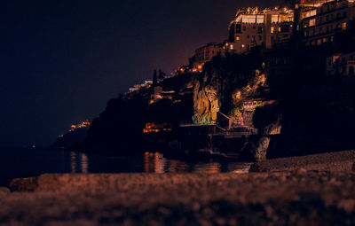 Illuminated buildings by sea against sky at night