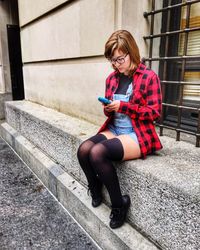 Young woman using mobile phone while sitting on retaining wall outside building