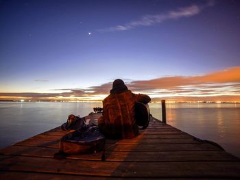 Rear view of man playing guitar while sitting on jetty over lake against sky at dusk