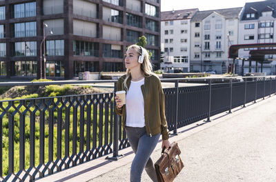 Young woman walking on bridge, drinking coffee, listening music with headphones