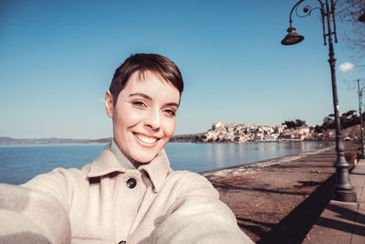The young woman with short hair is taking a selfie in front of the lake.