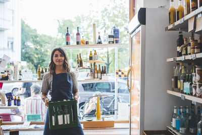 Portrait of confident female employee carrying bottles in crate at deli