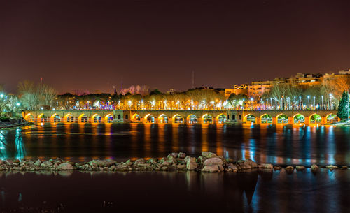 Illuminated bridge over river by buildings against sky at night