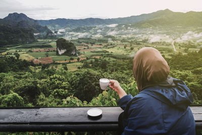 Rear view of woman drinking coffee on railing against mountains and sky