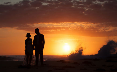 Silhouette couple standing at beach against sky during sunset