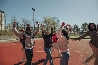 Carefree multiracial friends with arms raised dancing together at sports court on sunny day