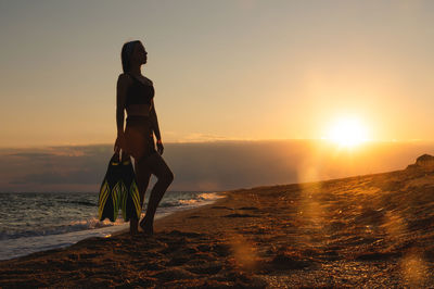 Rear view of woman standing on beach against sky during sunset