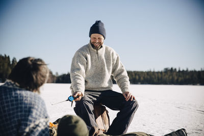 Happy mature man holding fishing rod sitting with friends during sunny day in winter