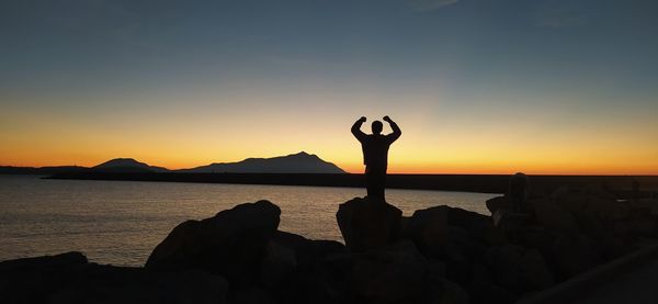 Silhouette man standing on rock by sea against sky during sunset