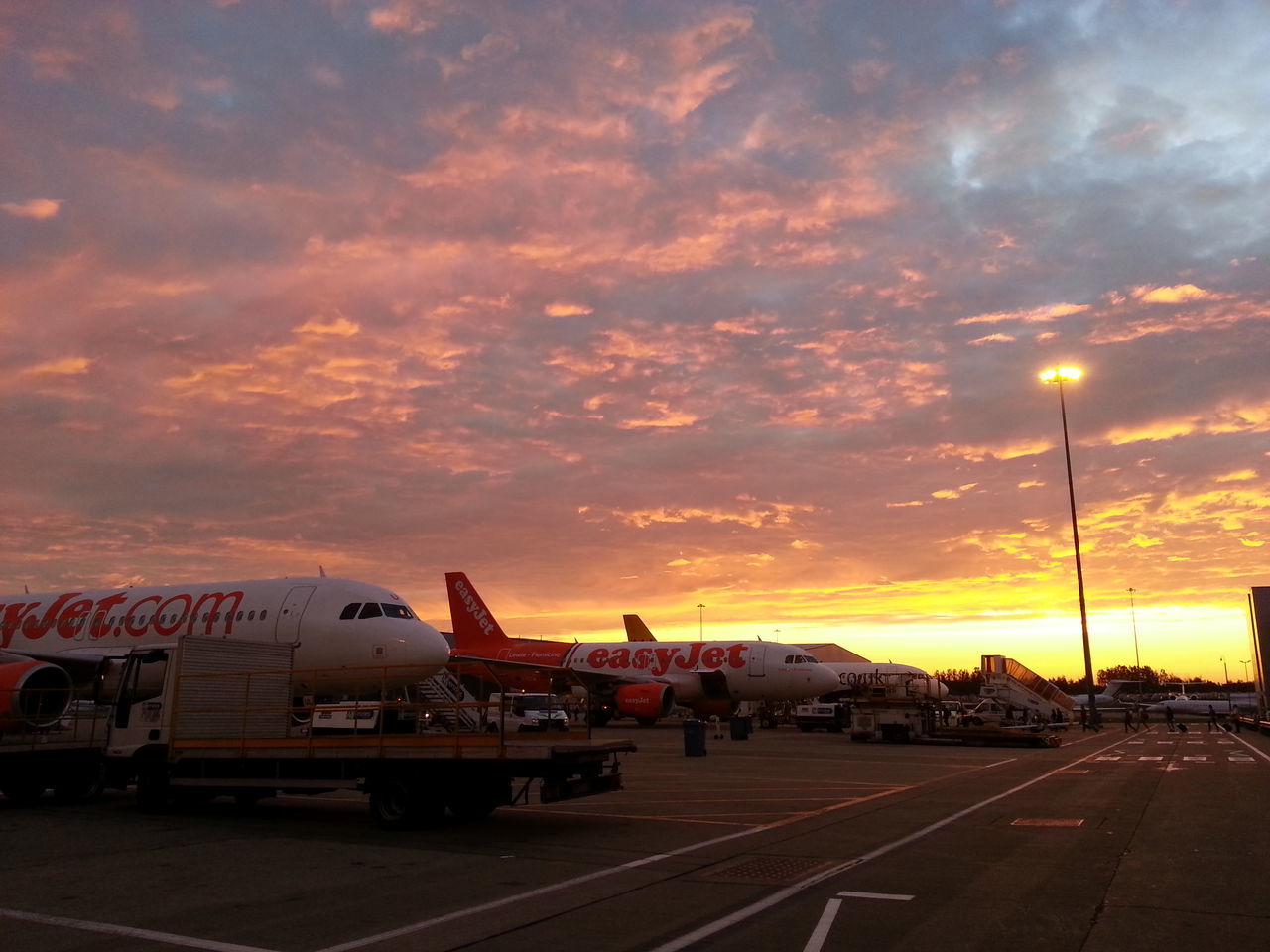 transportation, sky, sunset, cloud - sky, mode of transport, road, land vehicle, car, cloudy, road marking, street, the way forward, cloud, street light, orange color, outdoors, dusk, parking lot, dramatic sky, travel