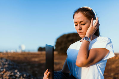 Young woman wearing headphones while using laptop against sky