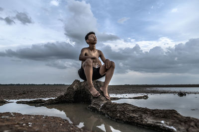 Shirtless boy sitting on rock against cloudy sky during arid climate