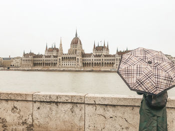 View of city by river against cloudy sky with woman standing with umbrella overlooking budapest parl
