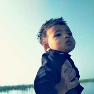 Cropped image of hand carrying baby boy by lake against clear sky