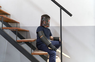 Cute boy wearing cardboard mask and holding sword sitting on staircase at home