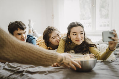 Hand of mother serving popcorn to children taking selfie through smart phone while lying down on bed at home