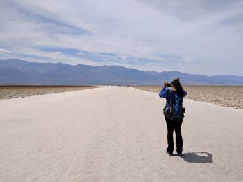 Rear view of woman photographing while standing on sand against cloudy sky
