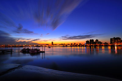 Illuminated city by sea against sky at sunset