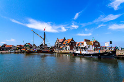 Sailboats in river by buildings against blue sky