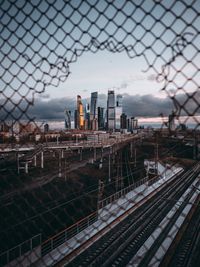 Railroad tracks seen through fence against cityscape during sunset