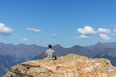 Young man from back meditating on rock against background of caucasus mountains and blue cloudy sky