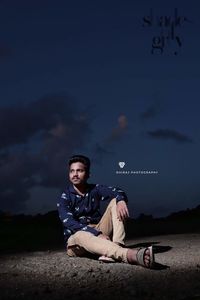 Young man looking away while sitting on land against sky at night