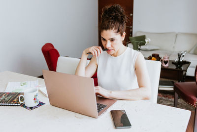 Focused female entrepreneur in casual clothes sitting at table with smartphone and notebooks and biting pen while working on netbook at home in daytime