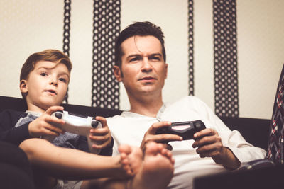 Father and son playing video games while sitting on sofa