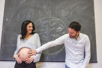 Smiling young man touching pregnant wife belly while standing by blackboard