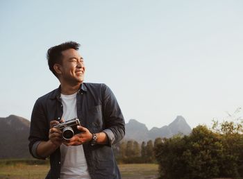 Smiling man holding camera against clear sky