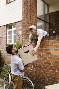 Man passing cardboard box to boyfriend while leaning on brick wall