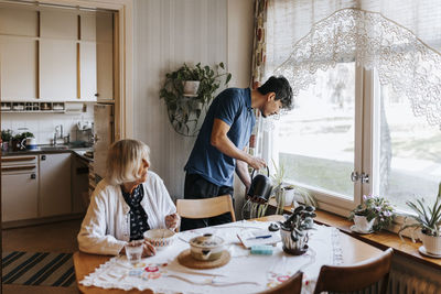 Male caregiver watering plants by senior woman eating food while sitting at dining table in home