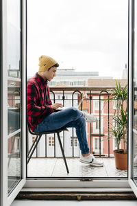 Side view of trendy focused caucasian man with beanie hat and shirt sitting on chair at balcony while working remotely on laptop at home