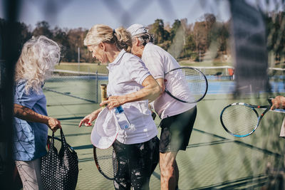 Senior male and female friends with rackets and bag talking while standing at tennis court