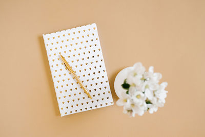 A white notebook with a gold pen on a beige table and flowers in a defocus vase. blogger's workplace