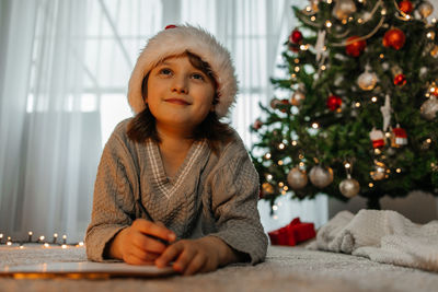 A girl in a red santa claus hat writes a letter to santa claus on the background of christmas tree