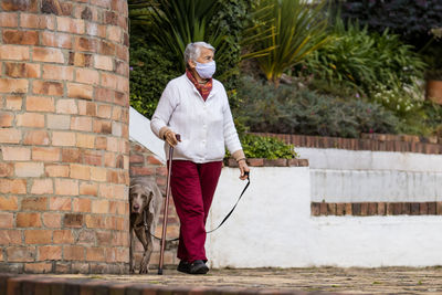 Full length of senior wearing mask with dog walking outdoors