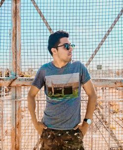 Young man wearing sunglasses standing against chainlink fence