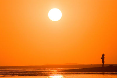 Silhouette woman standing on beach against orange sky