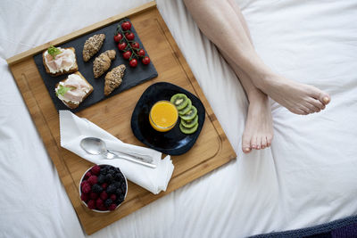 Close up of women's legs and a breakfast tray with fruits on a bed.