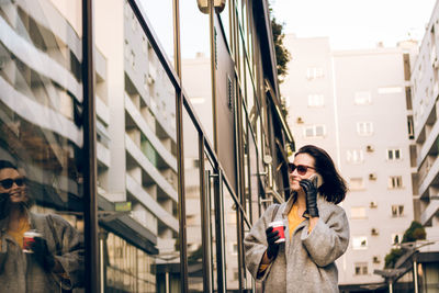 Woman drinking coffee while talking on phone in city during winter