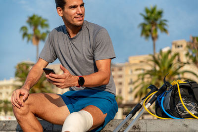 Young latin man with crutches using mobile phone after doing exercise