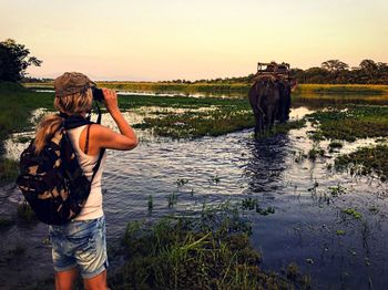 Woman looking through binoculars at lakeshore while elephant walking in background