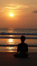 Man sitting on shore against orange sunset sky