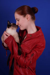 Young woman holding siamese cat against blue background