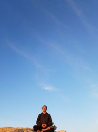 Low angle view of man meditating on cliff against blue sky