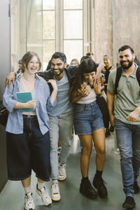 Happy man enjoying while walking with arm around female friends at college corridor