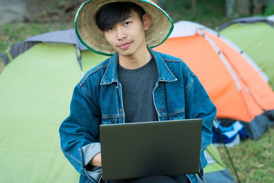 Portrait of young man with laptop sitting against tents