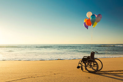 Colorful helium balloons on empty wheelchair at beach against clear blue sky