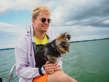 Handsome blond young person wear raincoat sitting on a sailing yacht and hold his small dog